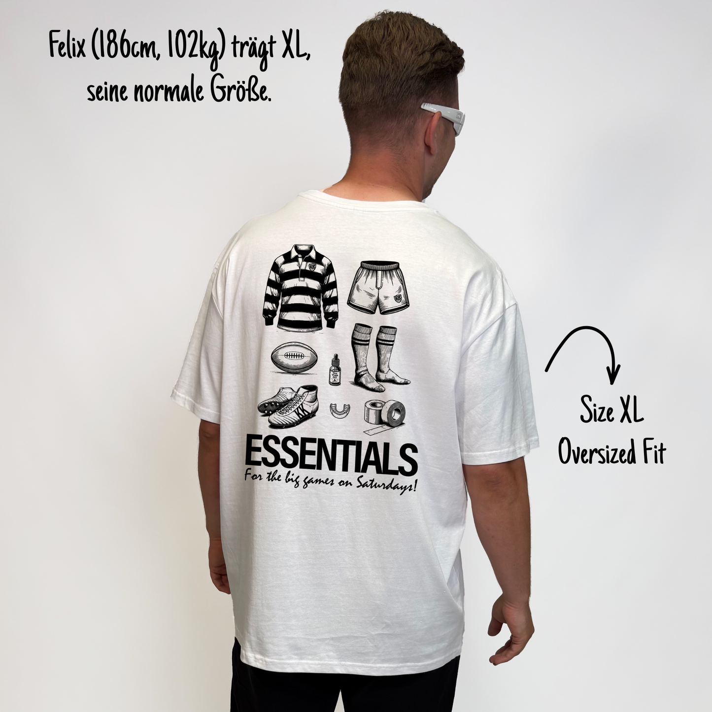 Felix (186cm, 102kg) trägt das weiße The Essentials Tee in XL. Man sieht das Model von hinten. Das Shirt ist oversized geschnitten und hat einen großen Print auf dem Rücken: ein Trikot, Shorts, ein Ball, Schuhe, Mundschutz, Tape, Teebaumöl und Stulpen. Untendrunter steht: "Essentials for the big games on Saturdays!"