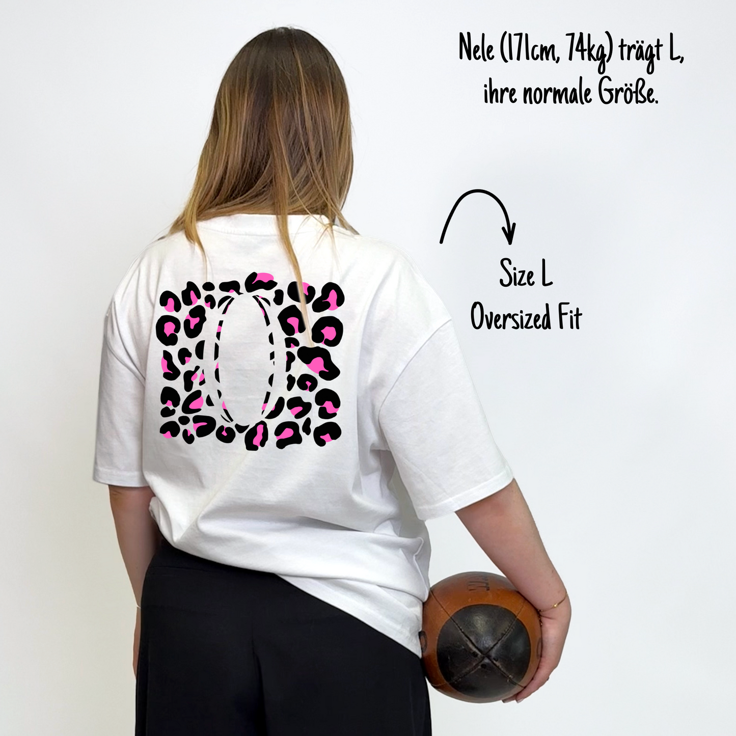 Nele (171cm, 74kg) trägt das weiße Klub Leo Tee in L. Man sieht das Model von hinten. Das Shirt ist oversized geschnitten und hat einen großen Print auf dem Rücken: pinke Leoparden-Flecken in einem Rechteck über den ganzen Rücken, in der Mitte ein großer Rugbyball.ausgespart.
