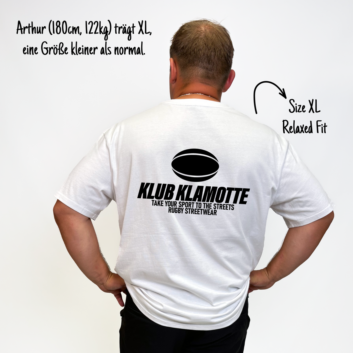 Arthur (180cm, 122kg) trägt das weiße Klub Streets Tee in XL. Man sieht das Model von hinten. Er trägt das Shirt im Relaxed Fit, eine Größe kleiner als normal. Das Shirt hat einen großen schwarzen Print auf dem Rücken: ein quer liegender Rugby Ball, darunter der Schriftzug Klub Klamotte und darunter "Take your sport to the streets, Rugby Streetwear".