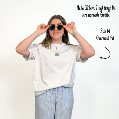 Naila (172cm, 71kg) trägt das weiße Klub Font Tee in M. Man sieht das Model von vorne. Sie trägt das Shirt im oversized Fit, ihre normale Größe. Das Shirt hat einen kleinen mittigen Print auf der Brust:  Ein grünes Rechteck mit dem weißen Schriftzug "KLUB" und darunter in schwarzer Schrift und durchgestrichen Klamotte.