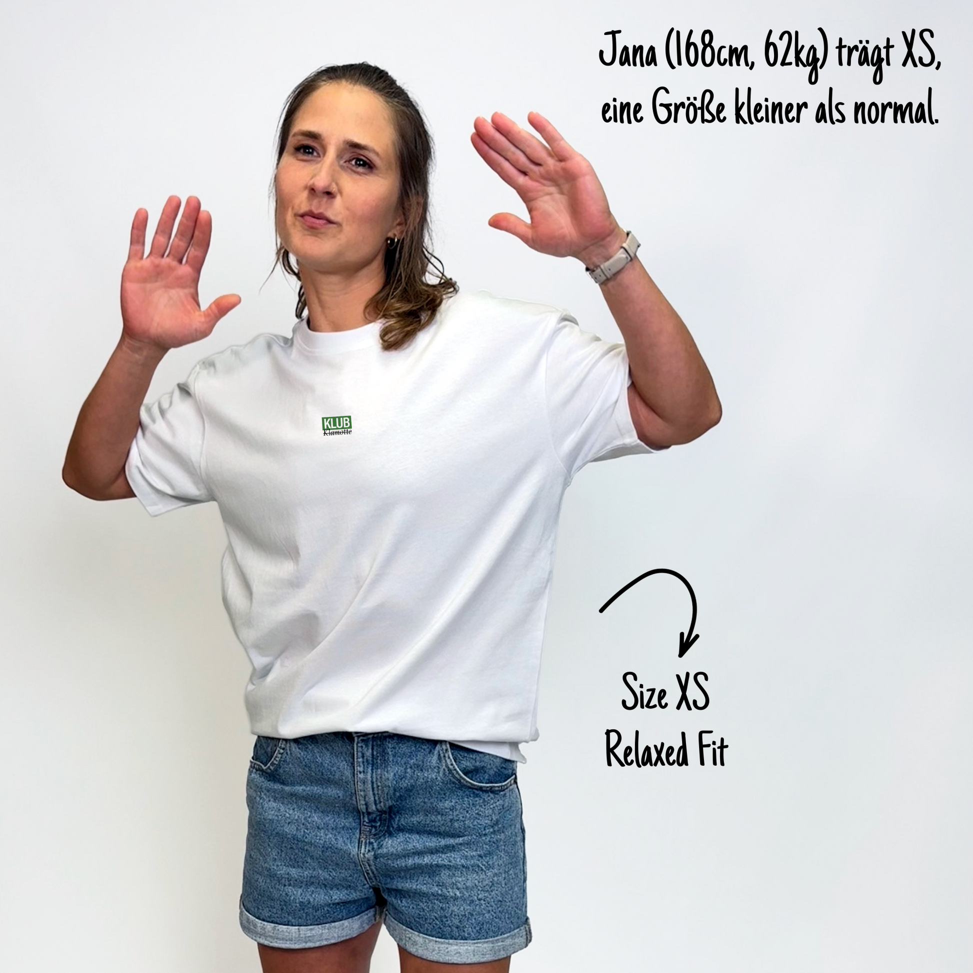Jana (168cm, 62kg) trägt das weiße Klub Font Tee in XS. Man sieht das Model von vorne. Sie trägt das Shirt im Relaxed Fit, eine Größe kleiner als normal. Das Shirt hat einen kleinen Print auf der Brust:  Ein grünes Rechteck mit dem weißen Schriftzug "KLUB" und darunter in schwarzer Schrift und durchgestrichen Klamotte.