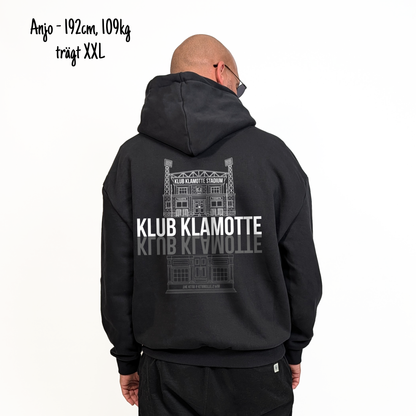 Anjo (192cm, 109kg) trägt den schwarzen From Klub to Pub Hoodie in XXL. Man sieht das Model von hinten. Der Hoodie hat einen großen Print auf dem Rücken: Ein Print des Klub Klamotte Stadium in weiß mit dem Schriftzug Klub Klamotte drunter. Darunter gespiegelt wieder der Schriftzug Klub Klamotte in grau und ein Print des Klub & Klamotte's Pub.