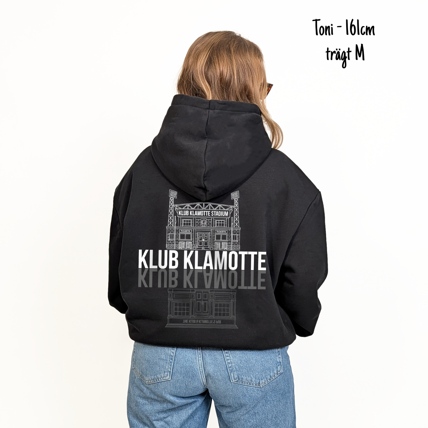 Toni (161cm) trägt den schwarzen From Klub to Pub Hoodie in M. Man sieht das Model von hinten. Der Hoodie hat einen großen Print auf dem Rücken: Ein Print des Klub Klamotte Stadium in weiß mit dem Schriftzug Klub Klamotte drunter. Darunter gespiegelt wieder der Schriftzug Klub Klamotte in grau und ein Print des Klub & Klamotte's Pub.