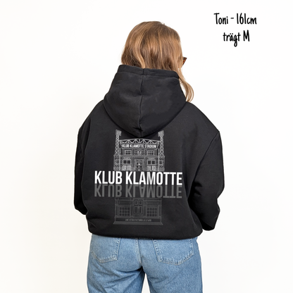 Toni (161cm) trägt den schwarzen From Klub to Pub Hoodie in M. Man sieht das Model von hinten. Der Hoodie hat einen großen Print auf dem Rücken: Ein Print des Klub Klamotte Stadium in weiß mit dem Schriftzug Klub Klamotte drunter. Darunter gespiegelt wieder der Schriftzug Klub Klamotte in grau und ein Print des Klub & Klamotte's Pub.