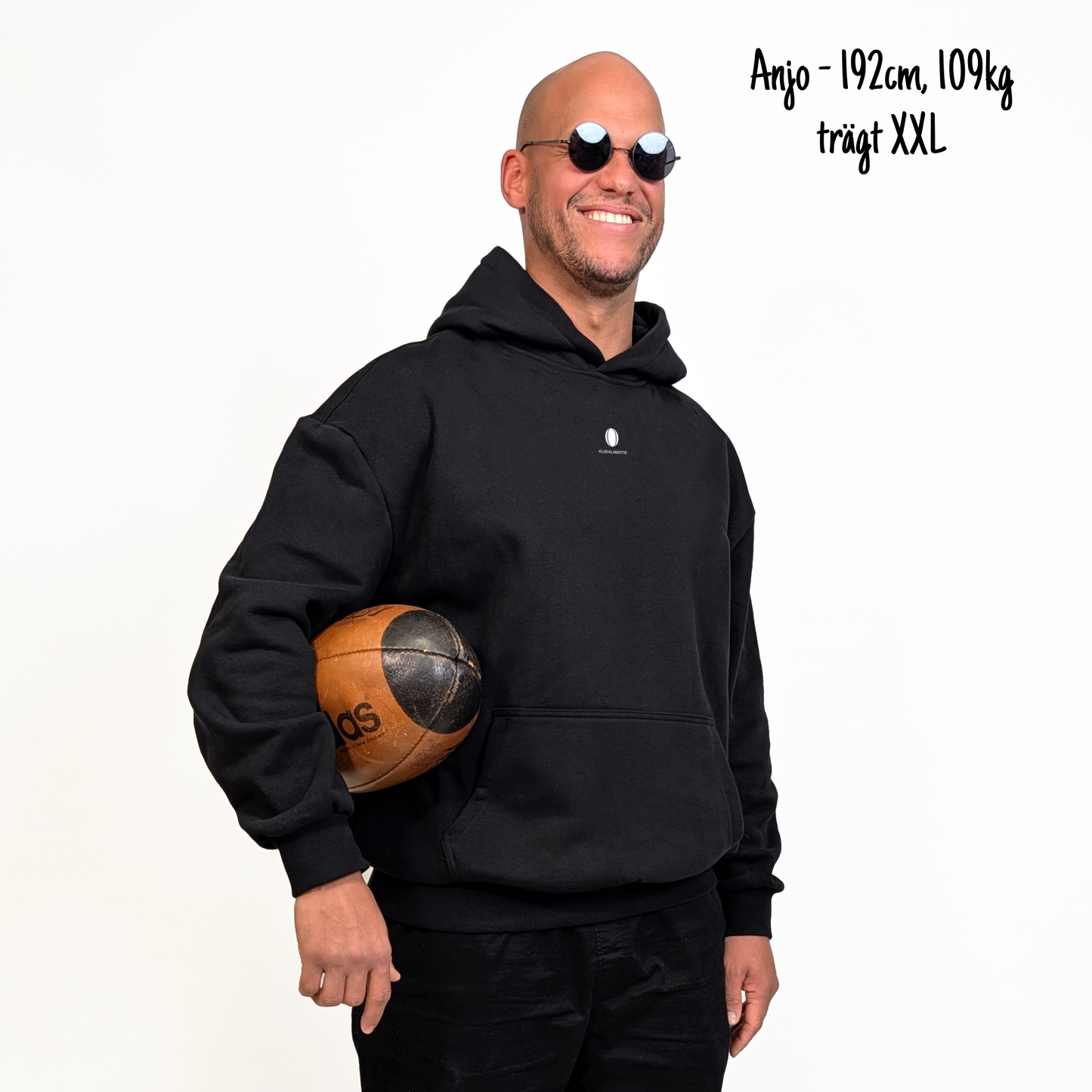 Anjo (192cm, 109kg) trägt den schwarzen Klub to Pub Hoodie in XXL. Man sieht das Model von vorne. Der Hoodie hat einen kleinen Print auf der Brust: das Klub Klamotte Logo.