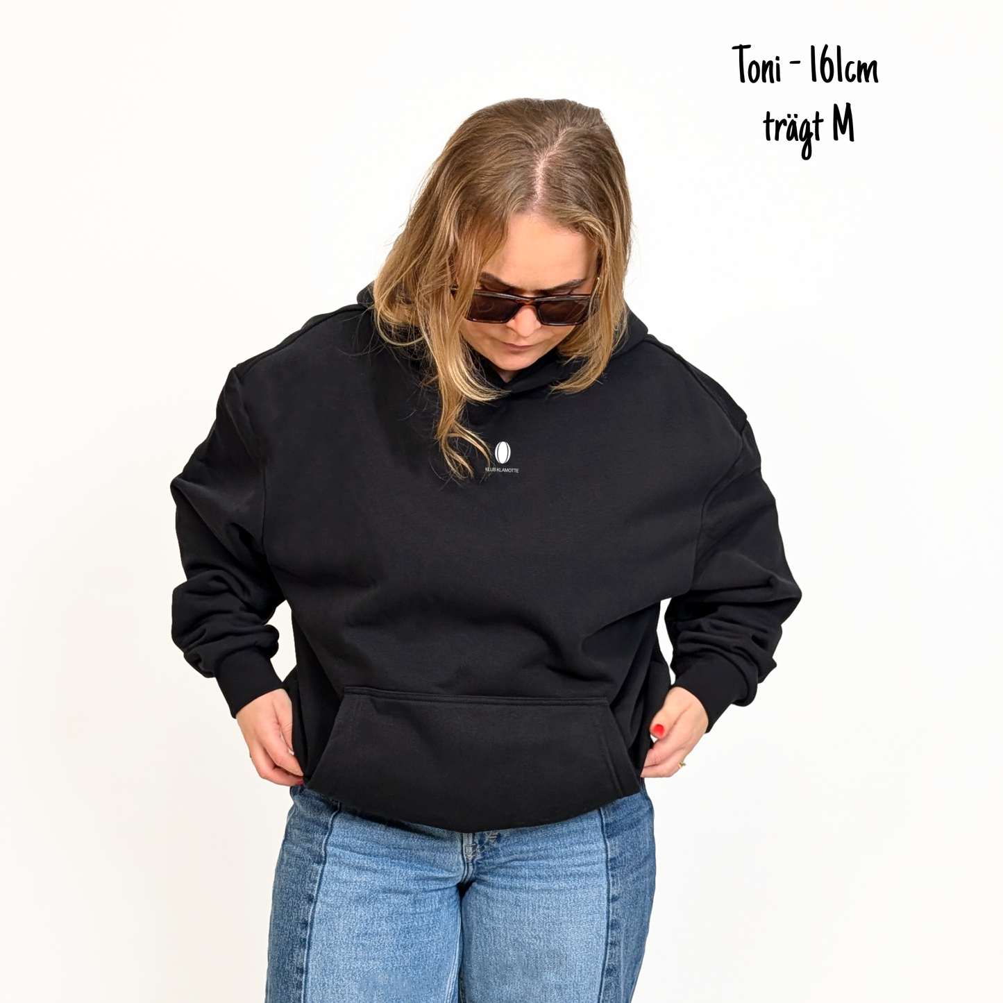 Toni (161cm) trägt den schwarzen Klub to Pub Hoodie in M. Man sieht das Model von vorne. Der Hoodie hat einen kleinen Print auf der Brust: das Klub Klamotte Logo.