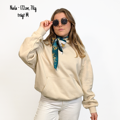 Naila (172cm, 71kg) trägt den sandfarbenen From Klub to Pub Hoodie in M. Man sieht das Model von vorne. Der Hoodie hat einen kleinen Print auf der Brust: das Klub Klamotte Logo. 