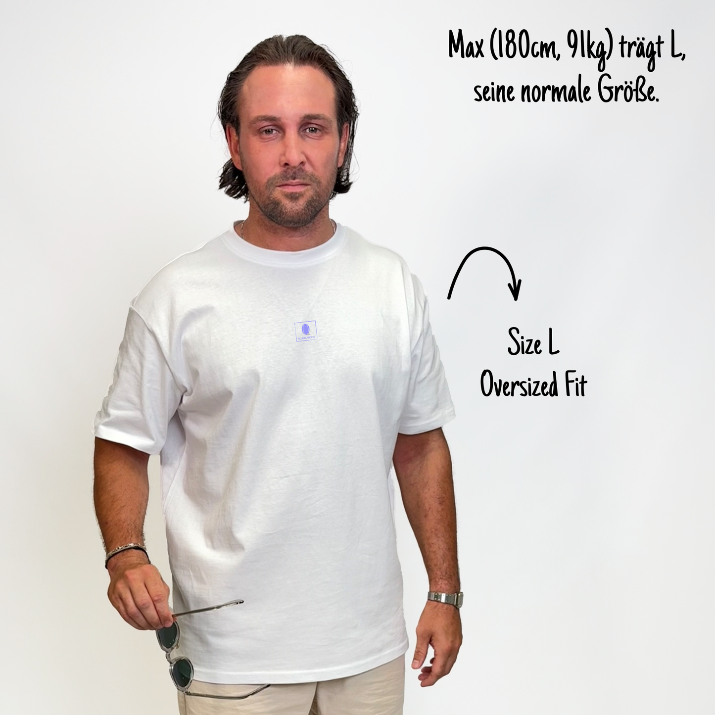 Max (180cm, 91kg) trägt das weiße Klub Print Tee in L. Man sieht das Model von vorne. Das Shirt ist oversized geschnitten und hat einen kleinen lilafarbenen Print auf der Brust: Das Klub Klamotte Logo in einem Quadrat.