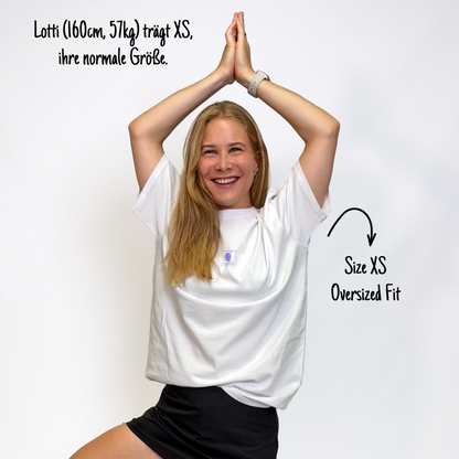 Lotti (160cm, 57kg) trägt das weiße Klub Print Tee in XS. Man sieht das Model von vorne. Das Shirt ist oversized geschnitten und hat einen kleinen lilafarbenen Print auf der Brust: Das Klub Klamotte Logo in einem Quadrat.
