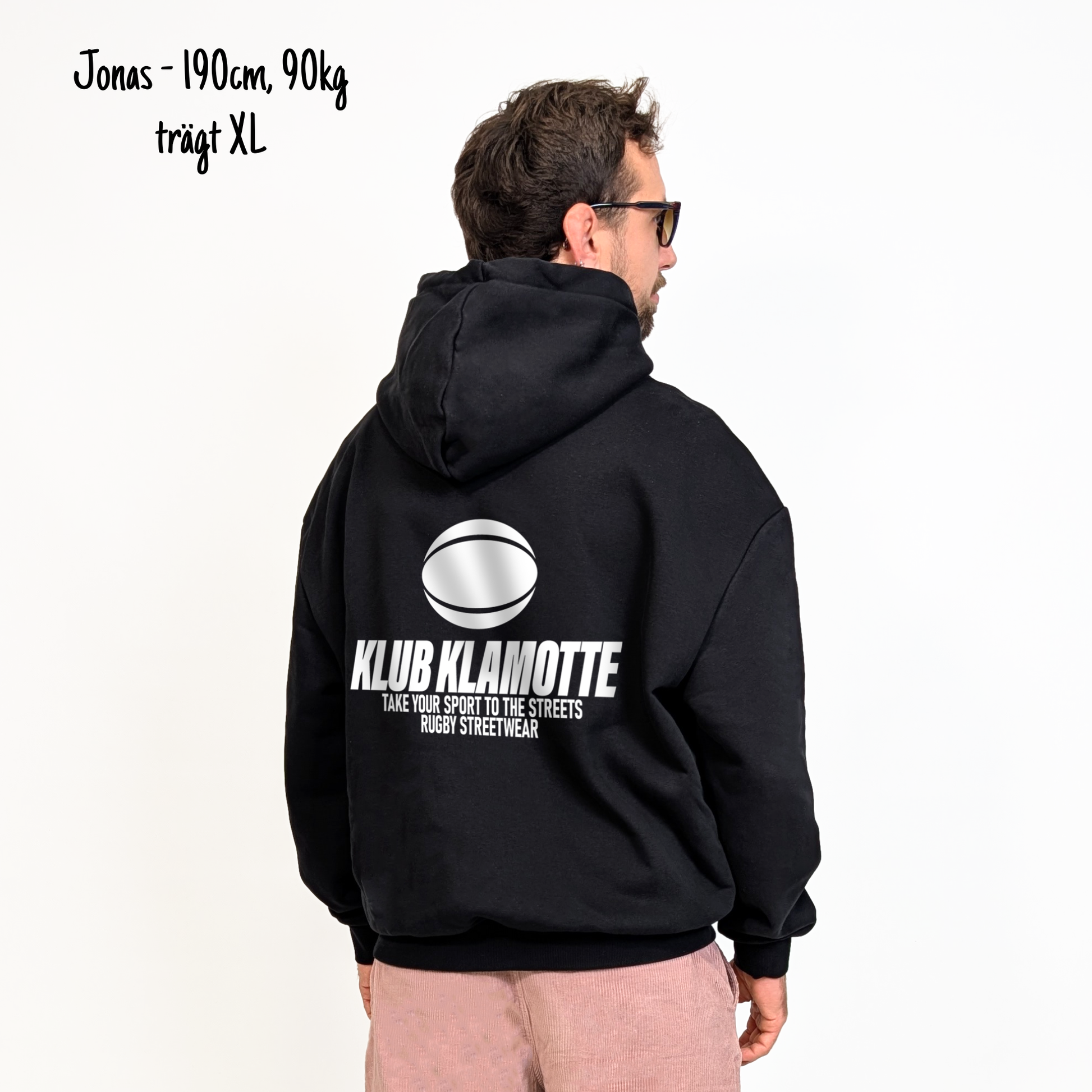 Jonas (190cm, 90kg) trägt den schwarzen Klub Streets Hoodie in XL. Man sieht das Model von hinten. Der Hoodie hat einen großen Print auf dem Rücken: ein quer liegender Rugby Ball, darunter der Schriftzug Klub Klamotte und darunter "Take your sport to the streets, Rugby Streetwear".