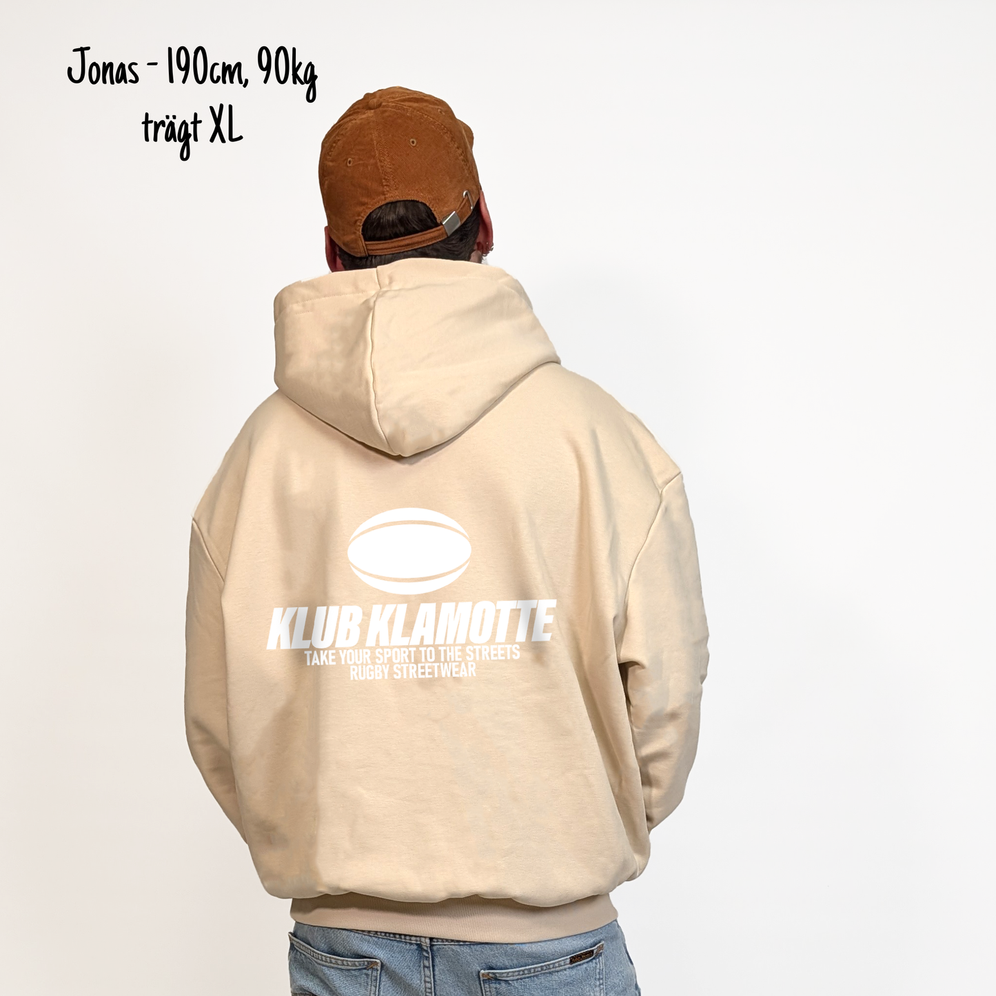 Jonas (190cm, 90kg) trägt den sandfarbenen Klub Streets Hoodie in XL. Man sieht das Model von hinten. Der Hoodie hat einen großen Print auf dem Rücken: ein quer liegender Rugby Ball, darunter der Schriftzug Klub Klamotte und darunter "Take your sport to the streets, Rugby Streetwear".