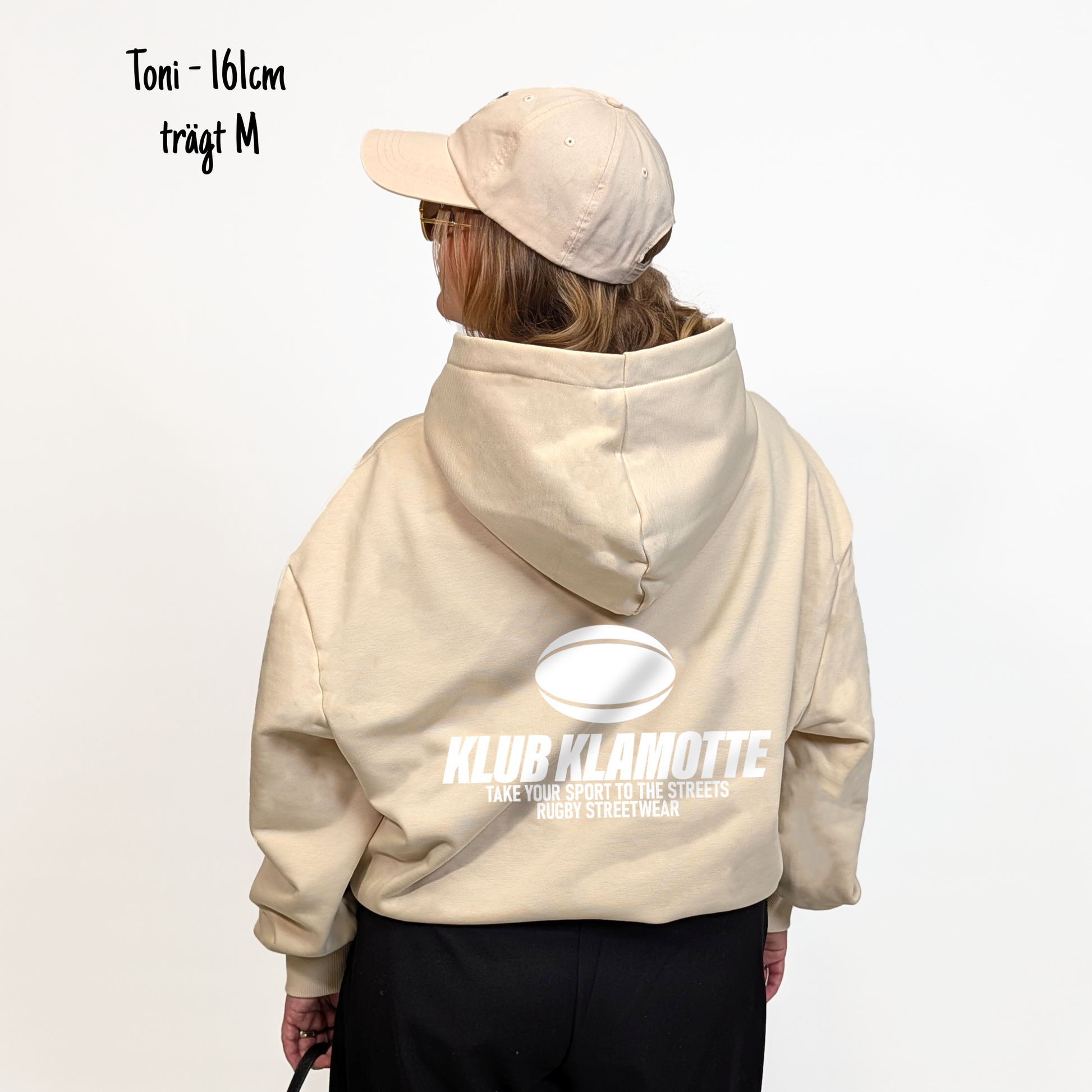 Toni (161cm) trägt den sandfarbenen Klub Streets Hoodie in M. Man sieht das Model von hinten. Der Hoodie hat einen großen Print auf dem Rücken: ein quer liegender Rugby Ball, darunter der Schriftzug Klub Klamotte und darunter "Take your sport to the streets, Rugby Streetwear".