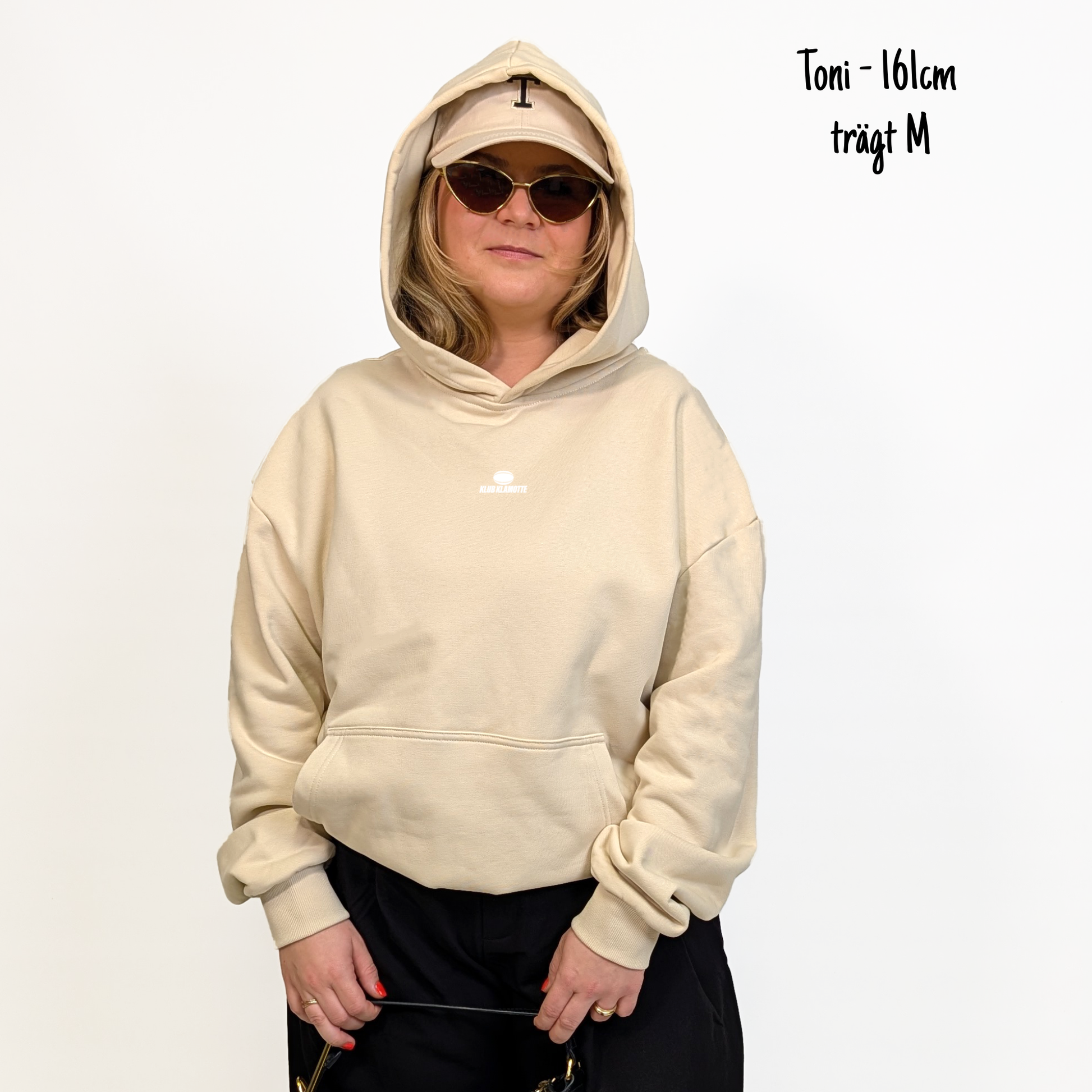 Toni (161cm) trägt den sandfarbenen Klub Streets Hoodie in M. Man sieht das Model von vorne. Der Hoodie hat einen kleinen Print auf der Brust: ein quer liegender Rugby Ball, darunter der Schriftzug Klub Klamotte.