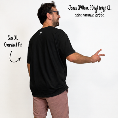 Jonas (190cm, 90kg) trägt das schwarze Klub Tee in XL. Man sieht das Model von hinten. Das Shirt ist oversized geschnitten und hat einen Print im Nacken: Das weiße Klub Klamotte Logo.