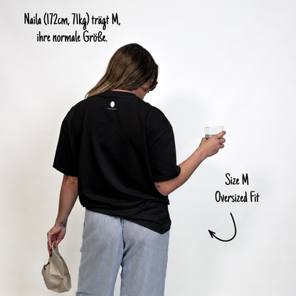 Naila (172cm, 71kg) trägt das schwarze Klub Tee in XL. Man sieht das Model von hinten. Das Shirt ist oversized geschnitten und hat einen Print im Nacken: Das weiße Klub Klamotte Logo.