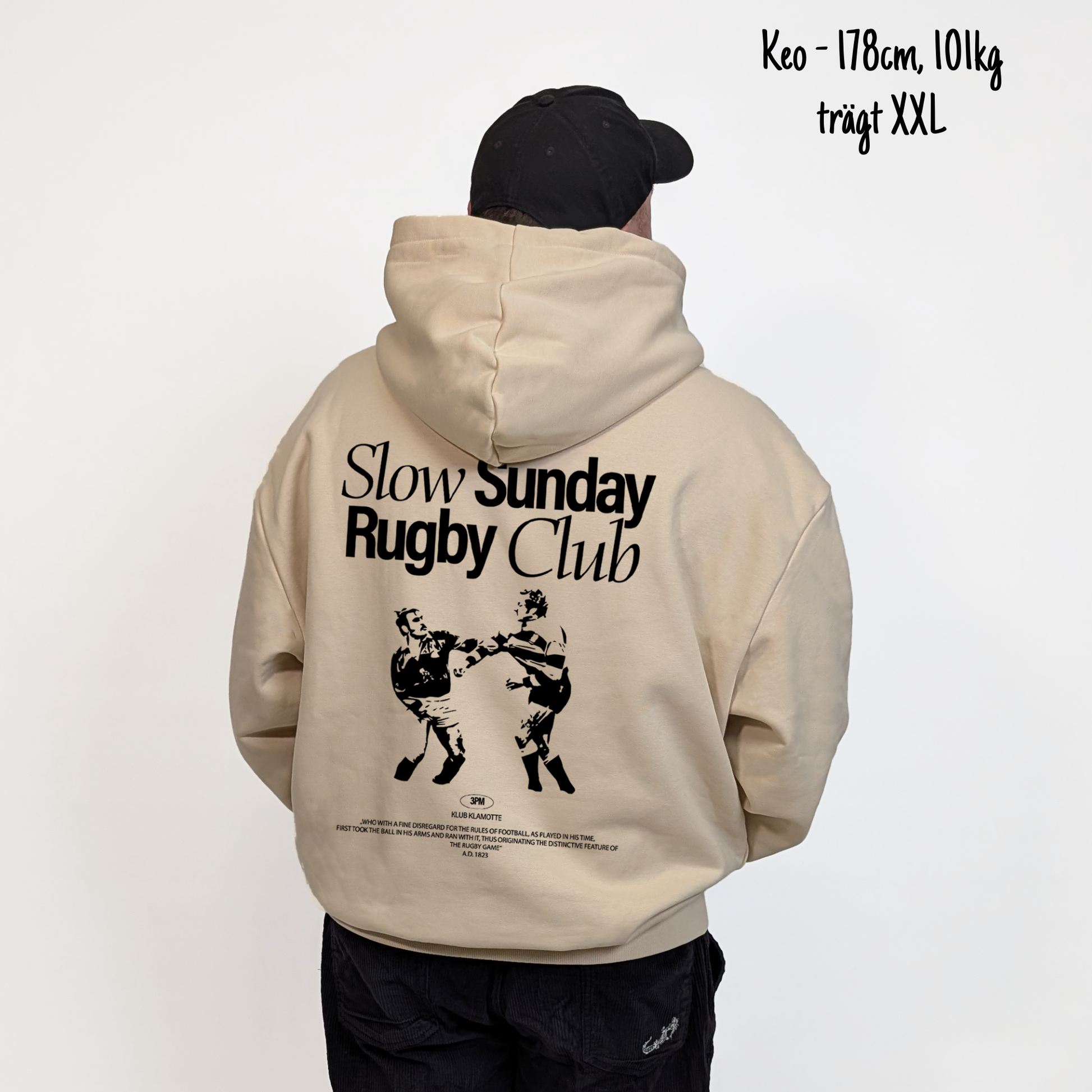 Keo (178cm, 101kg) trägt den sandfarbenen Slow Sunday Rugby Club Hoodie in XXL. Man sieht das Model von hinten. Der Hoodie hat einen großen Print auf dem Rücken: der Schriftzug Slow Sunday Rugby Club Tee und zwei Rugbyspieler im Streit. 