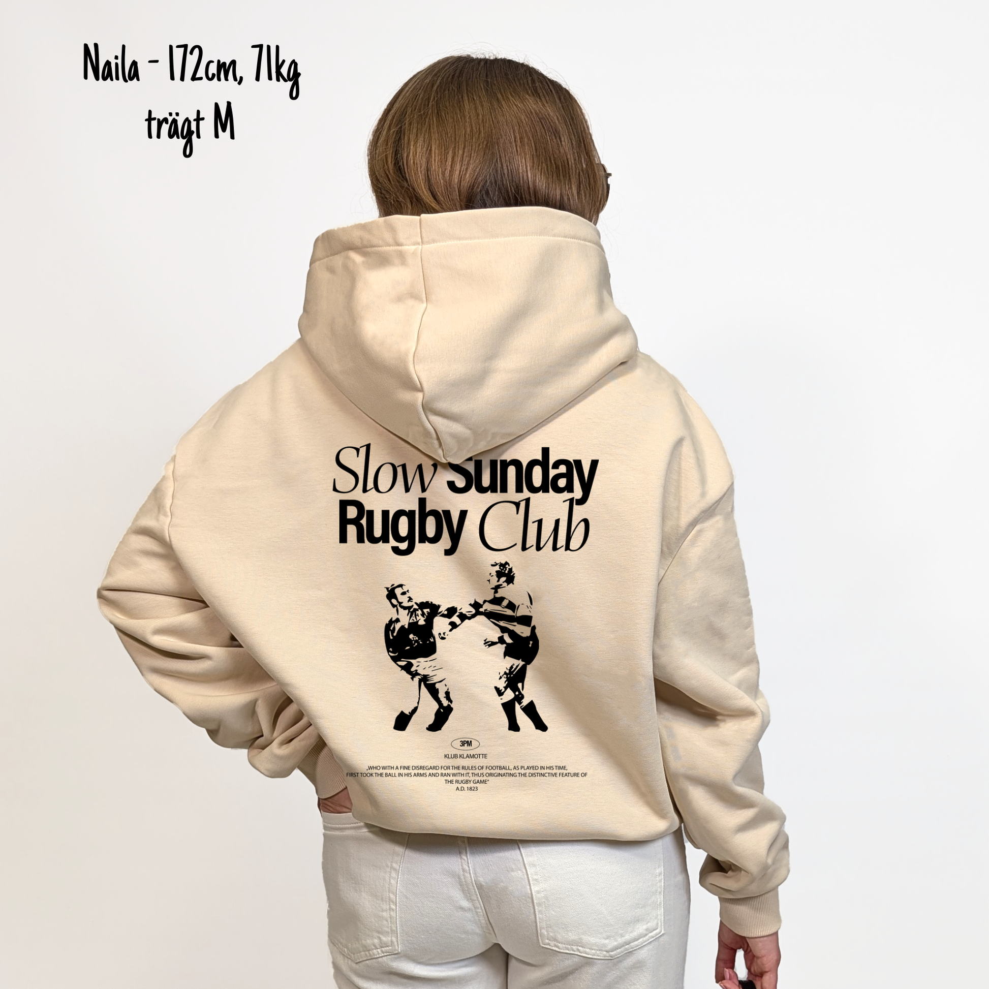 Naila (172cm, 71kg) trägt den sandfarbenen Slow Sunday Rugby Club Hoodie in M. Man sieht das Model von hinten. Der Hoodie hat einen großen Print auf dem Rücken: der Schriftzug Slow Sunday Rugby Club Tee und zwei Rugbyspieler im Streit. 