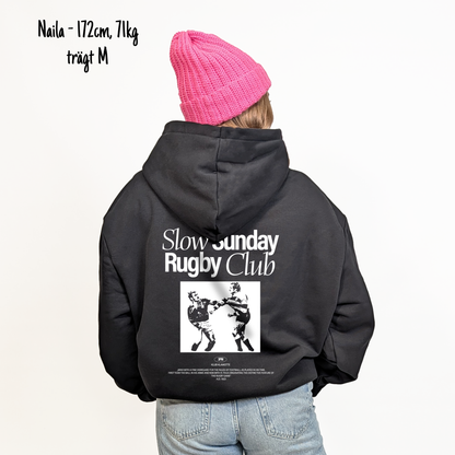 Naila (172cm, 71kg) trägt den schwarzen Slow Sunday Rugby Club Hoodie in M. Man sieht das Model von hinten. Der Hoodie hat einen großen Print auf dem Rücken: der Schriftzug Slow Sunday Rugby Club Tee und zwei Rugbyspieler im Streit. 