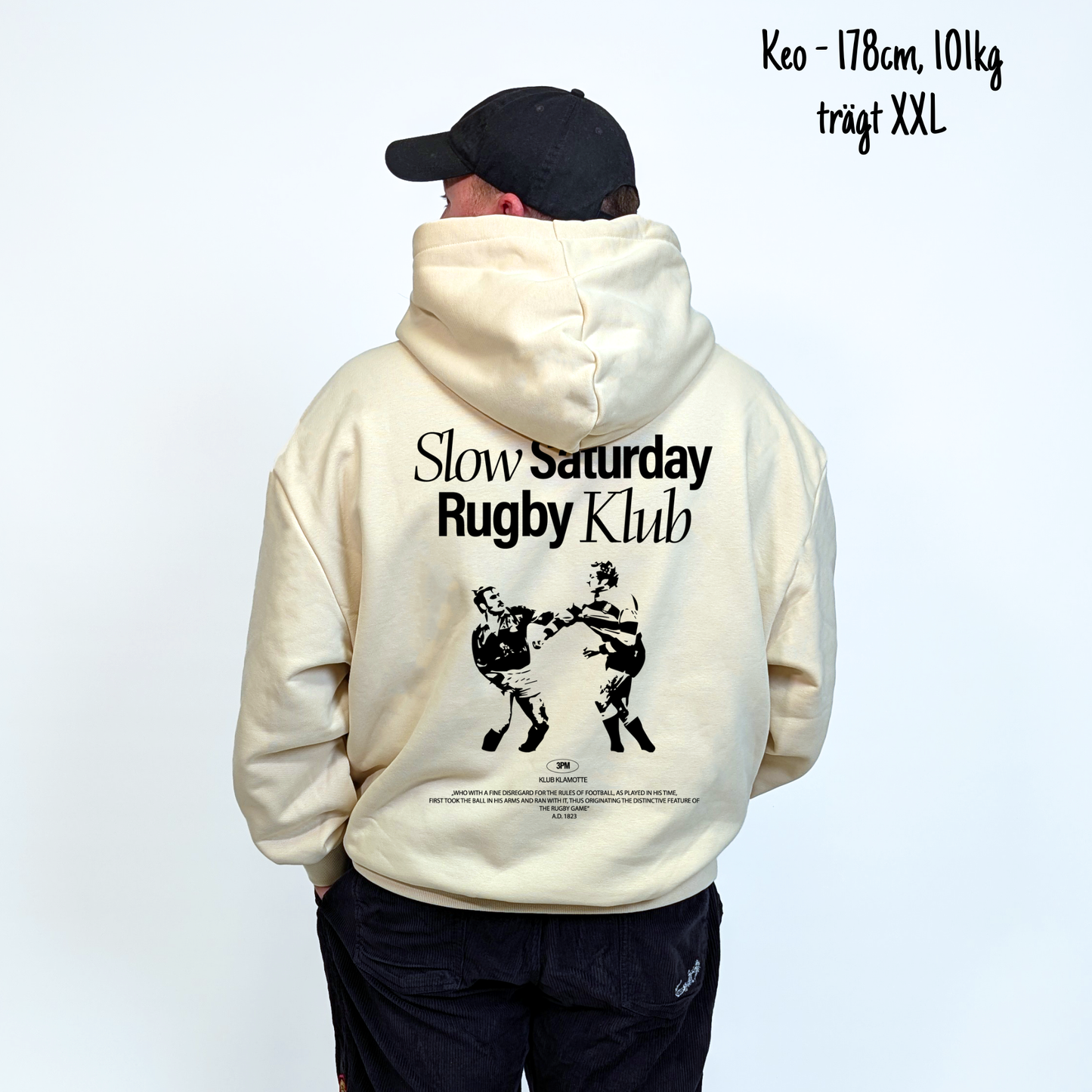 Keo (178cm, 101kg) trägt den sandfarbenen Slow Sunday Rugby Klub Hoodie in XXL. Man sieht das Model von hinten. Der Hoodie hat einen großen Print auf dem Rücken: der Schriftzug Slow Sunday Rugby Klub Tee und zwei Rugbyspieler im Streit. 