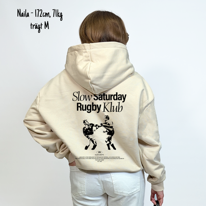 Naila (172cm, 71kg) trägt den sandfarbenen Slow Sunday Rugby Klub Hoodie in M. Man sieht das Model von hinten. Der Hoodie hat einen großen Print auf dem Rücken: der Schriftzug Slow Sunday Rugby Klub Tee und zwei Rugbyspieler im Streit. 
