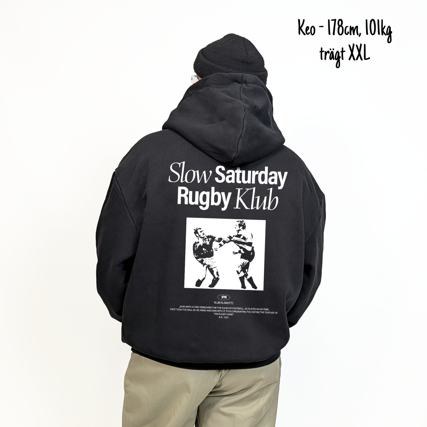 Keo (178cm, 101kg) trägt den schwarzen Slow Sunday Rugby Klub Hoodie in XXL. Man sieht das Model von hinten. Der Hoodie hat einen großen Print auf dem Rücken: der Schriftzug Slow Sunday Rugby Klub Tee und zwei Rugbyspieler im Streit. 