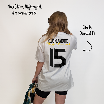 Naila (172 cm, 71 kg) trägt das weiße Team XV Tee in Größe M. Das Model ist von hinten zu sehen. Das Shirt ist oversized geschnitten und hat einen großen schwarzen Rückendruck mit der Nummer 15 in Tape-Optik. Darüber steht „Klub Klamotte“ in Schwarz und „Rugby Streetwear“ in Gold.