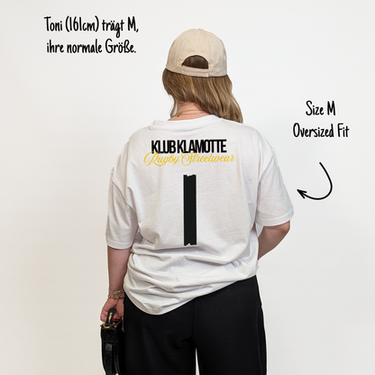 Toni (161 cm) trägt das weiße Team XV Tee in Größe M. Das Model ist von hinten zu sehen. Das Shirt ist oversized geschnitten und hat einen großen schwarzen Rückendruck mit der Nummer 1 in Tape-Optik. Darüber steht „Klub Klamotte“ in Schwarz und „Rugby Streetwear“ in Gold.